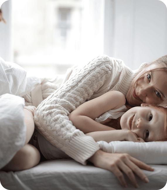 Une mère en pull en maille allongée à côté de sa fille dans un lit blanc pour un moment de câlin.