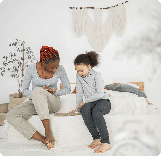 Une mère à la coiffure tressée rouge aidant sa fille lors d'une activité éducative sur un lit.